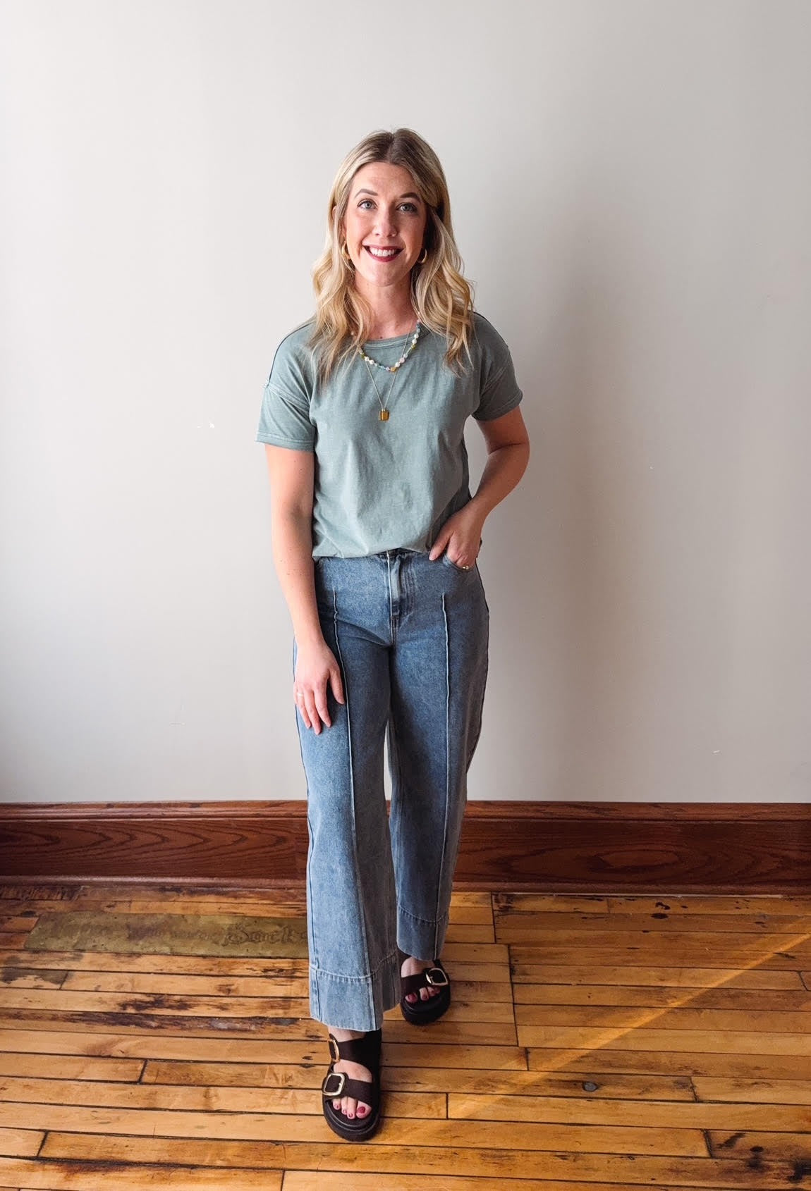 Woman wearing a green t-shirt and blue jeans standing on a wooden floor with a white wall background.