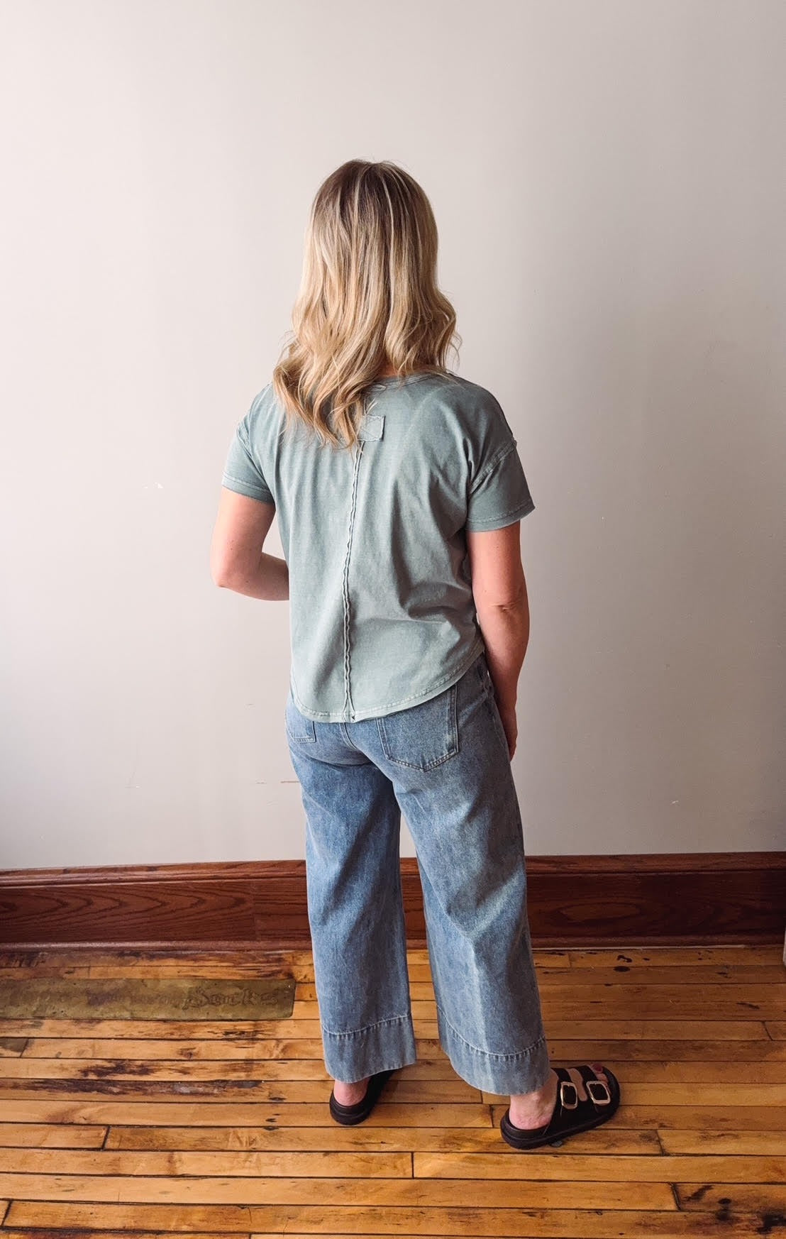 Person wearing a light green shirt and blue jeans standing on a wooden floor with a white wall background