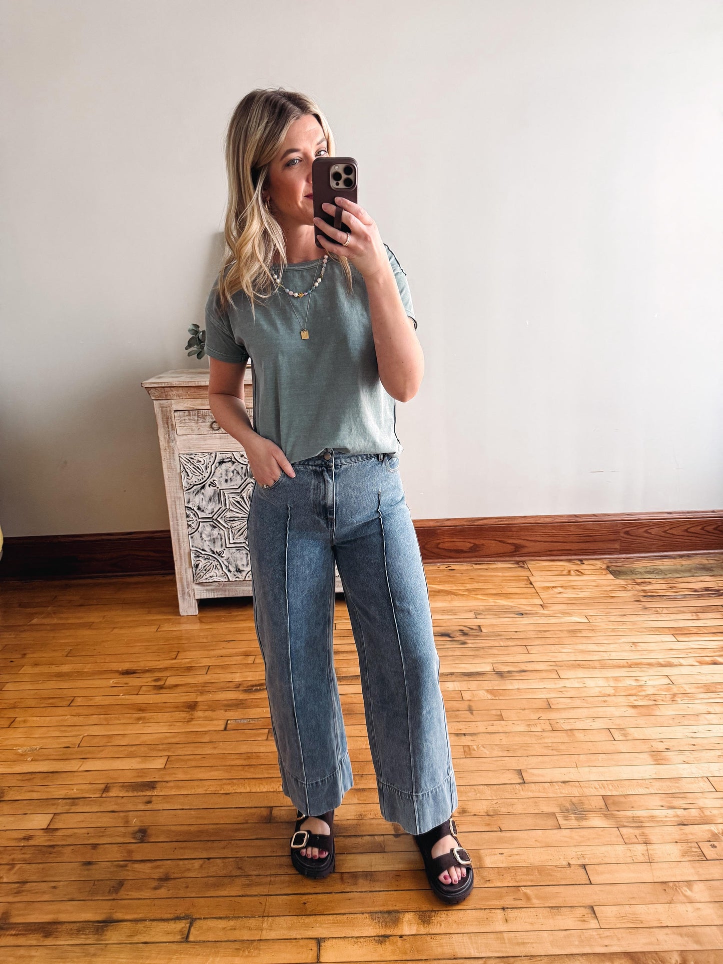 Woman taking a mirror selfie wearing a green t-shirt and blue jeans in a room with wooden flooring.