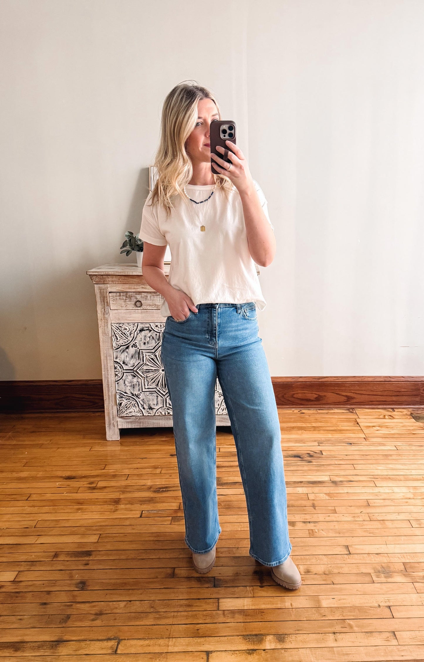 Woman taking a mirror selfie wearing a white t-shirt and blue jeans in a room with wooden flooring.