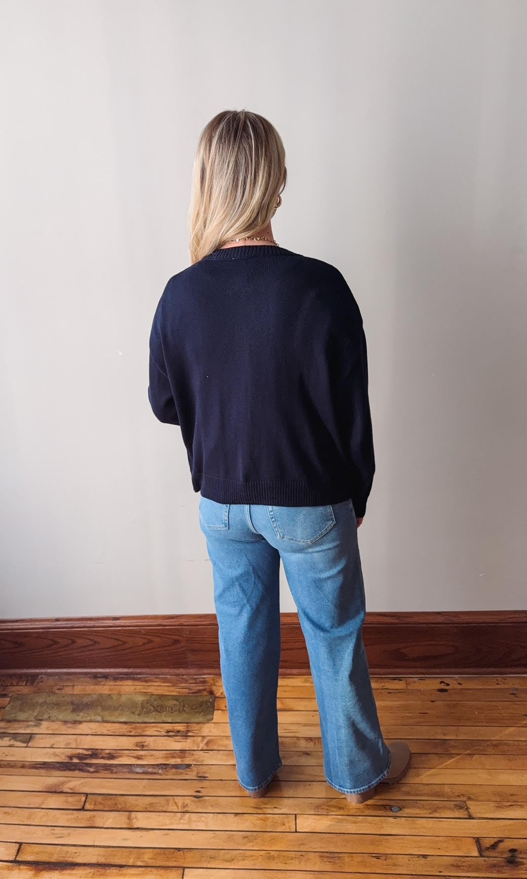 Person wearing a navy sweater and blue jeans standing in a room with wooden flooring and a white wall.