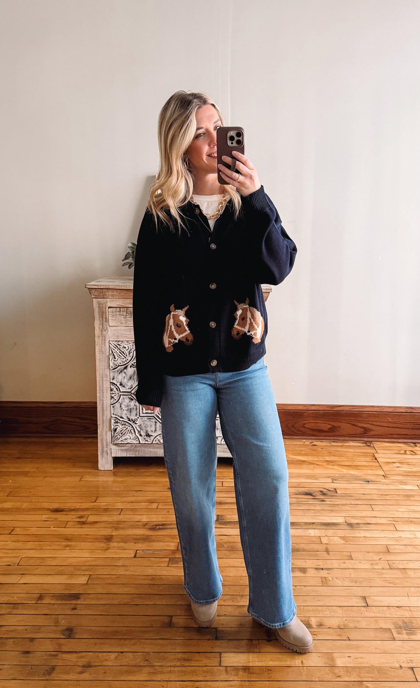 Woman taking a mirror selfie wearing a dark jacket and light blue jeans in a room with wooden flooring.