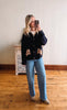 Woman taking a mirror selfie wearing a dark jacket and light blue jeans in a room with wooden flooring.