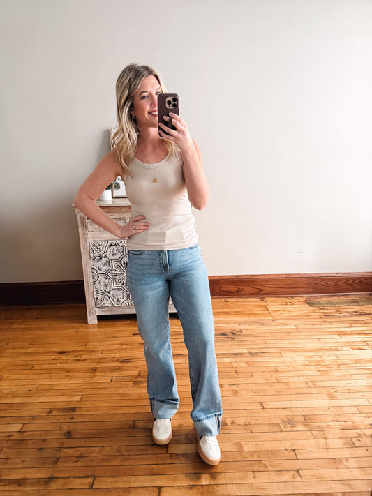 Woman taking a mirror selfie wearing a white tank top and blue jeans in a room with wooden flooring.