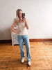 Woman taking a mirror selfie wearing a white tank top and blue jeans in a room with wooden flooring.