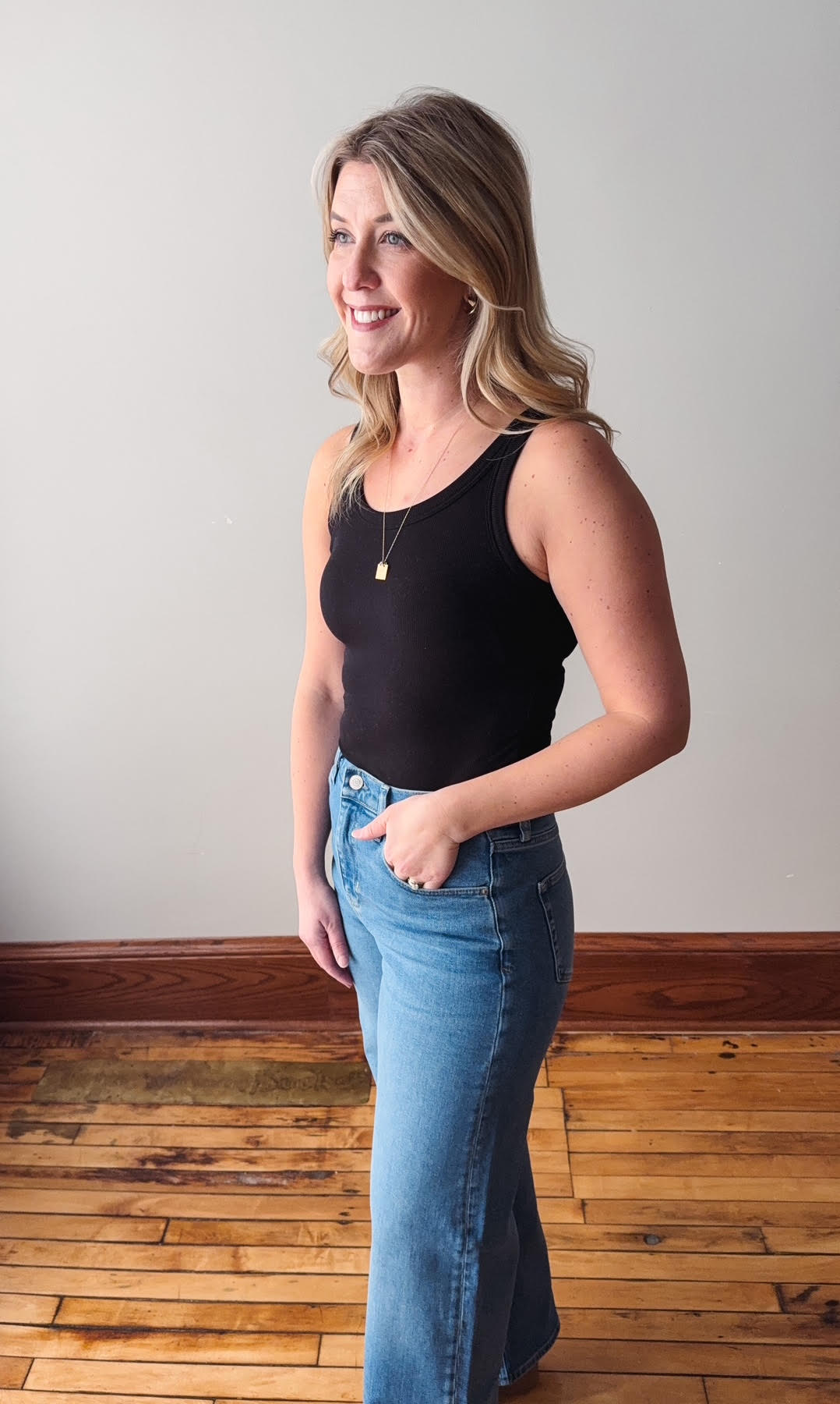 Woman wearing a black tank top and blue jeans standing on a wooden floor with a plain wall background