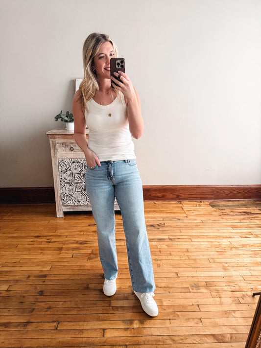 Woman taking a mirror selfie wearing a white tank top and light blue jeans in a room with wooden flooring.