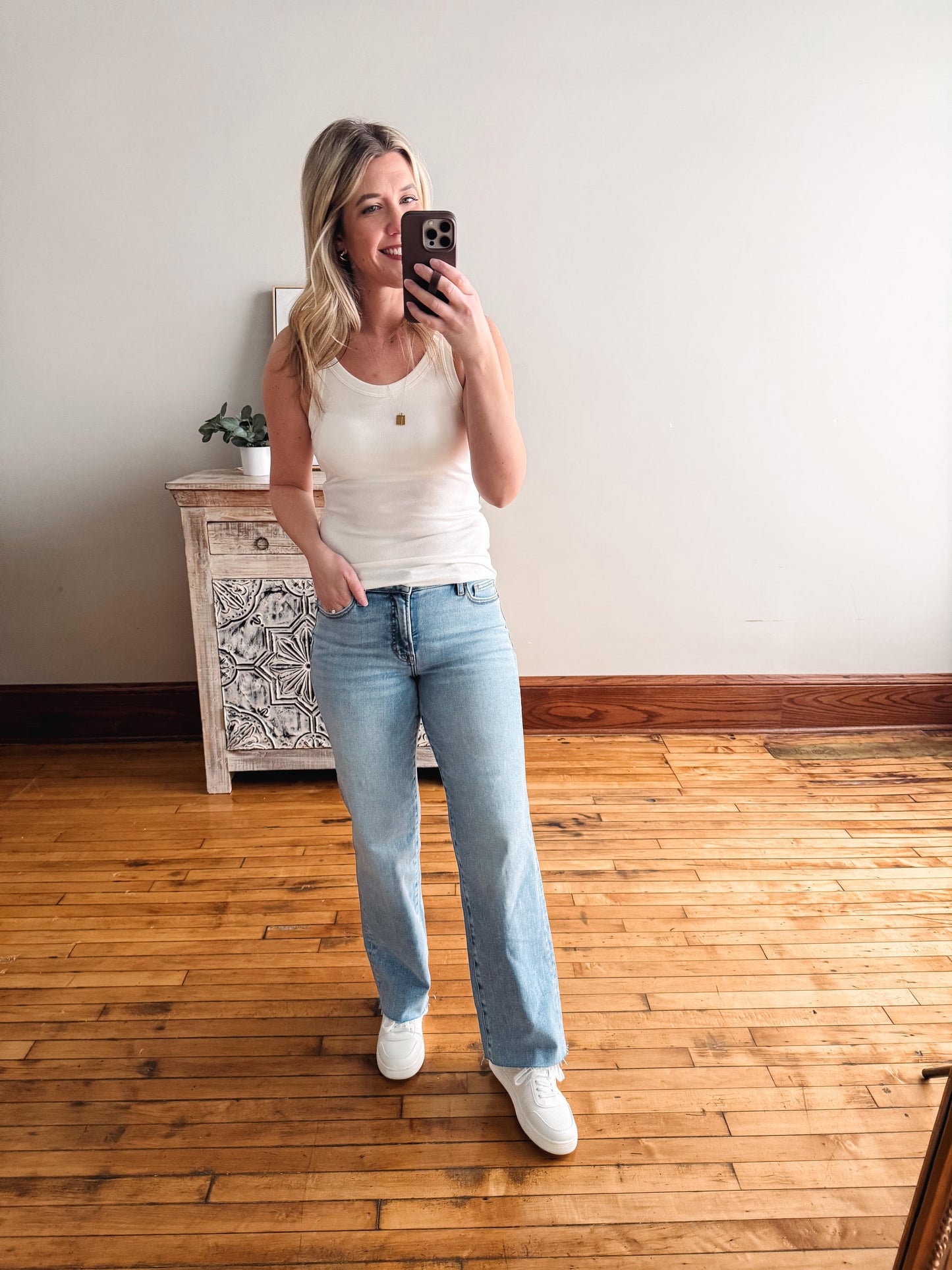 Woman taking a mirror selfie wearing a white tank top and light blue jeans in a room with wooden flooring.