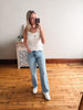 Woman taking a mirror selfie wearing a white tank top and light blue jeans in a room with wooden flooring.