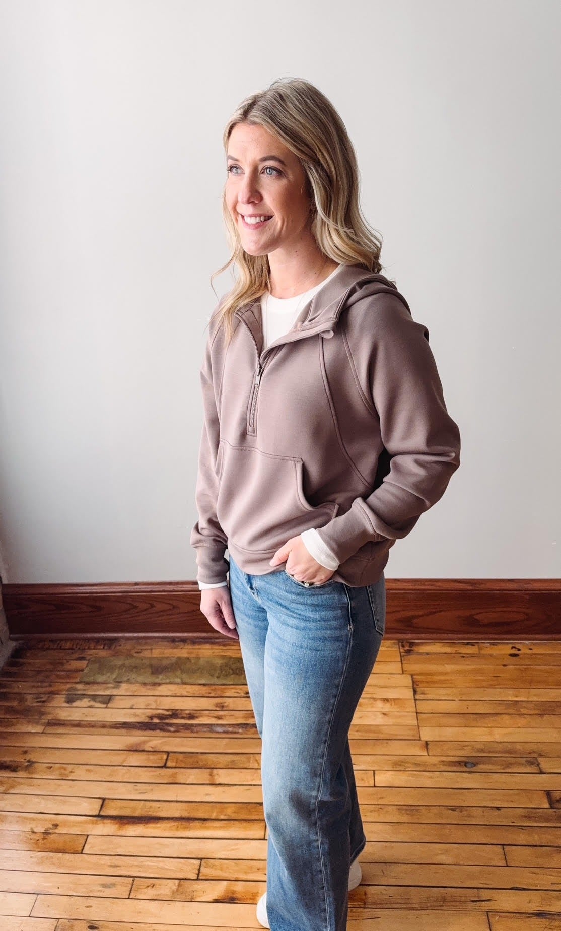 Woman wearing a brown hoodie and blue jeans standing on a wooden floor.