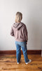 Person wearing a pink hoodie and blue jeans standing on a wooden floor with a white wall background
