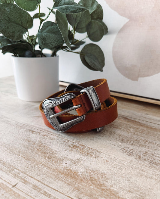 Brown leather belt with silver buckle on a wooden surface with a plant and decorative item in the background.