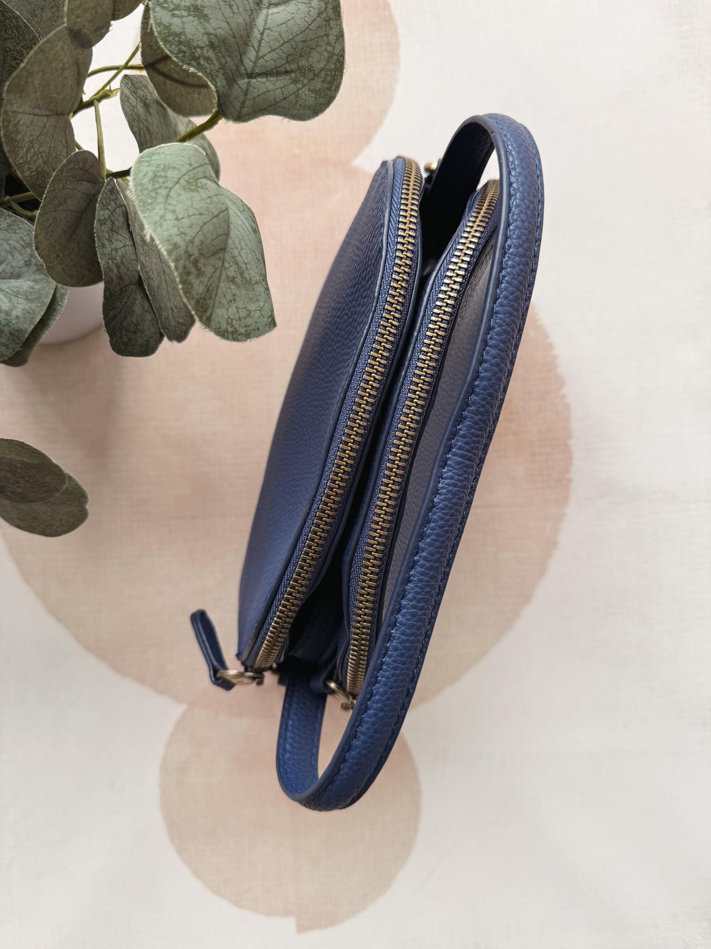 Blue leather clutch with gold zipper on a light pink background with green leaves.
