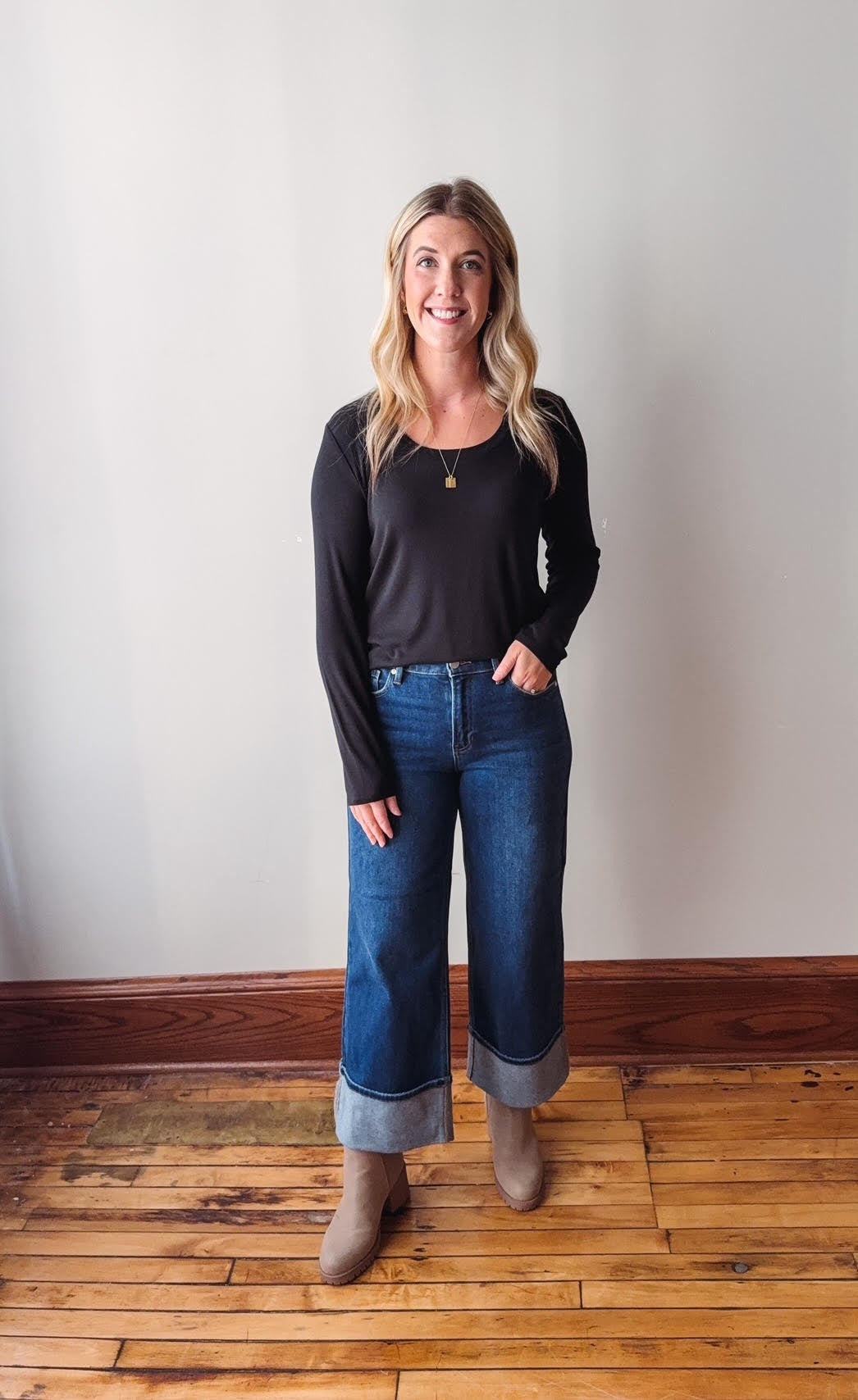 Woman wearing a black top and blue jeans standing on a wooden floor with a white wall background