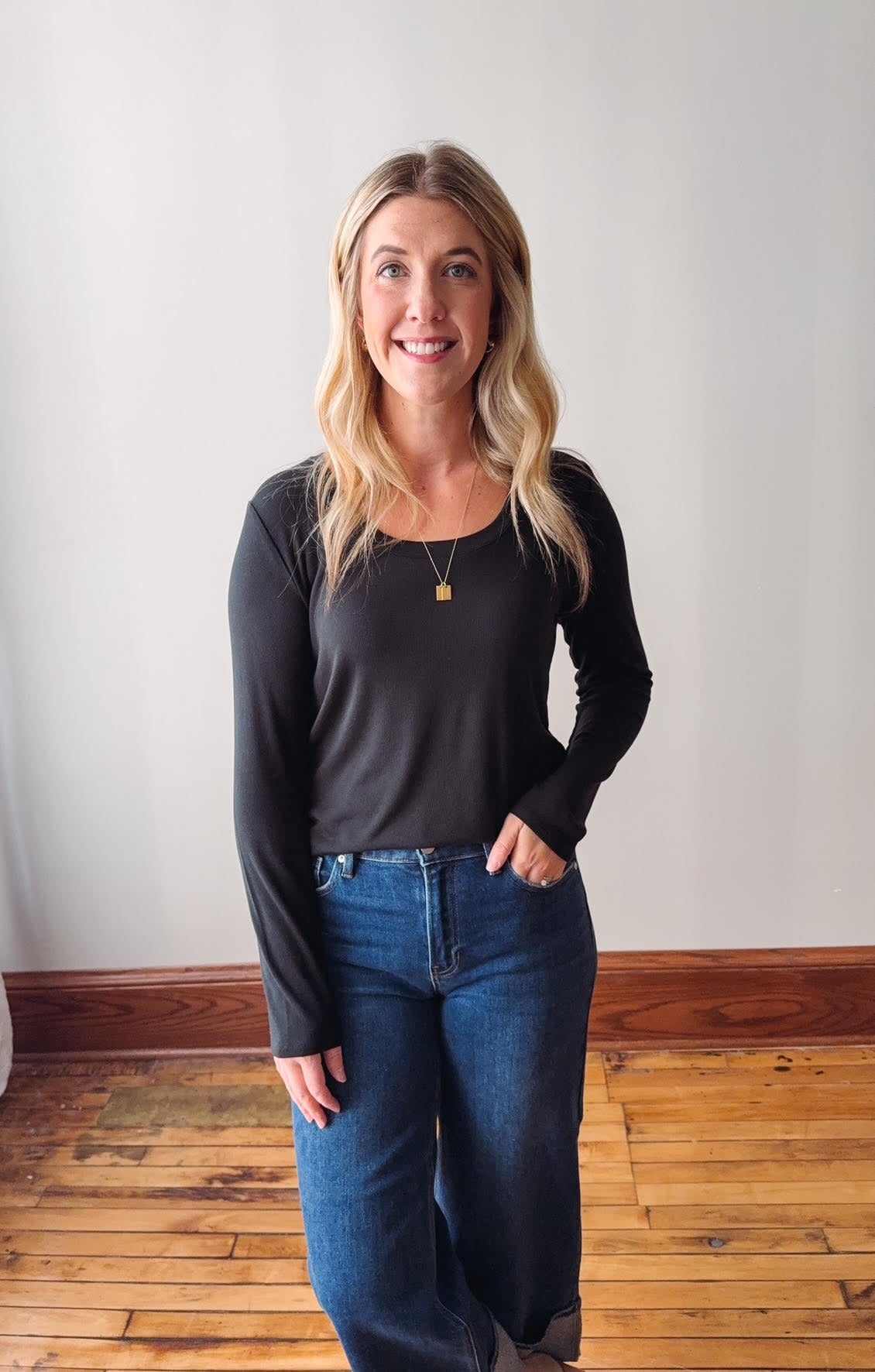 Woman wearing a black long-sleeve top and blue jeans standing on a wooden floor with a white wall background.