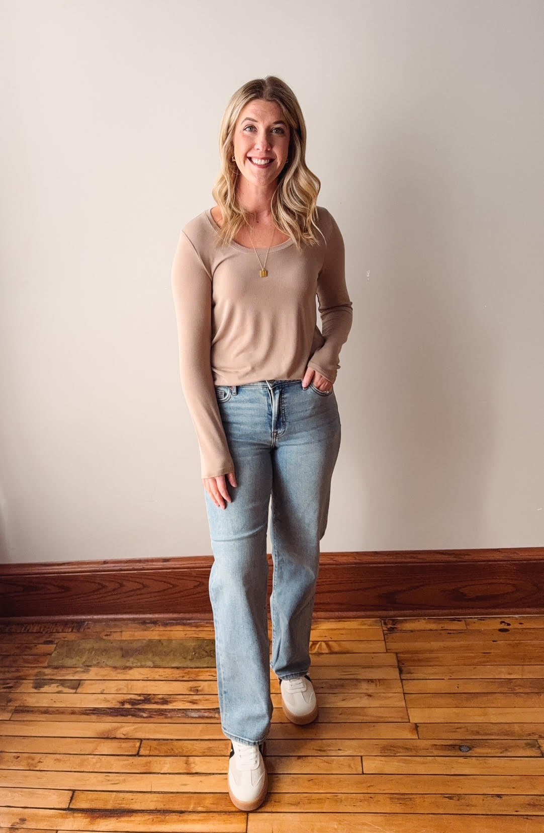 Woman wearing a beige top and light blue jeans standing on a wooden floor against a white wall.