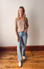 Woman wearing a beige top and light blue jeans standing on a wooden floor against a white wall.