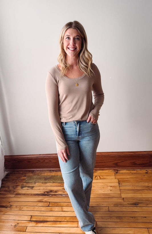 Woman wearing a beige long-sleeve top and light blue jeans standing on a wooden floor.