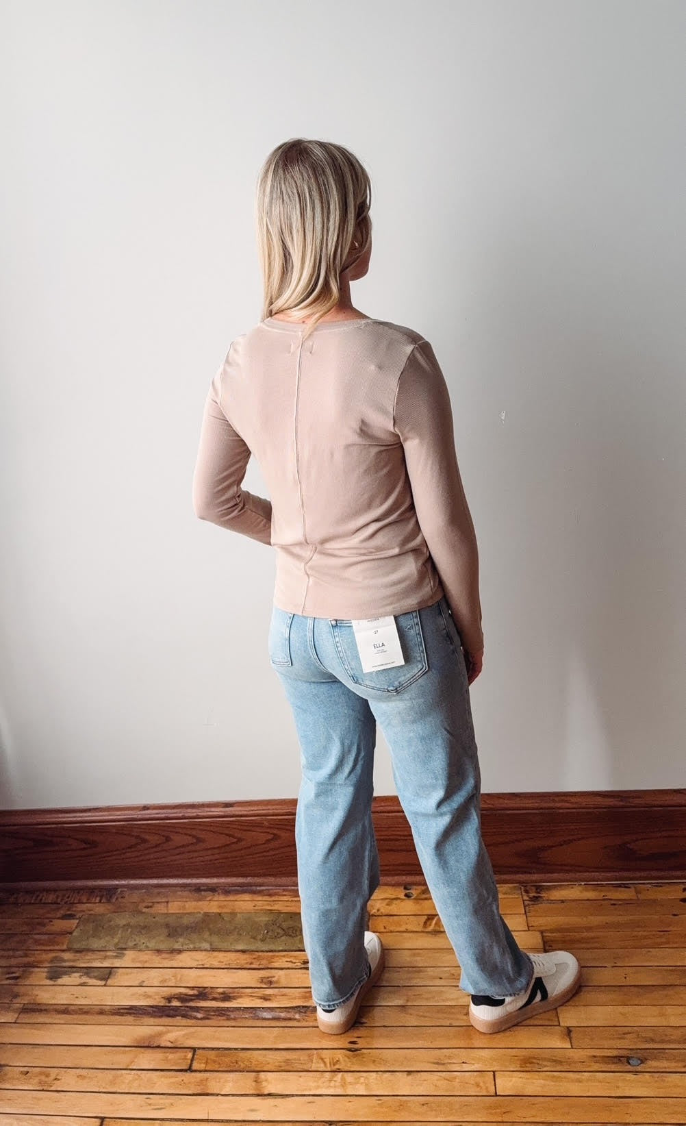 Person wearing a pink shirt and blue jeans standing against a white wall.