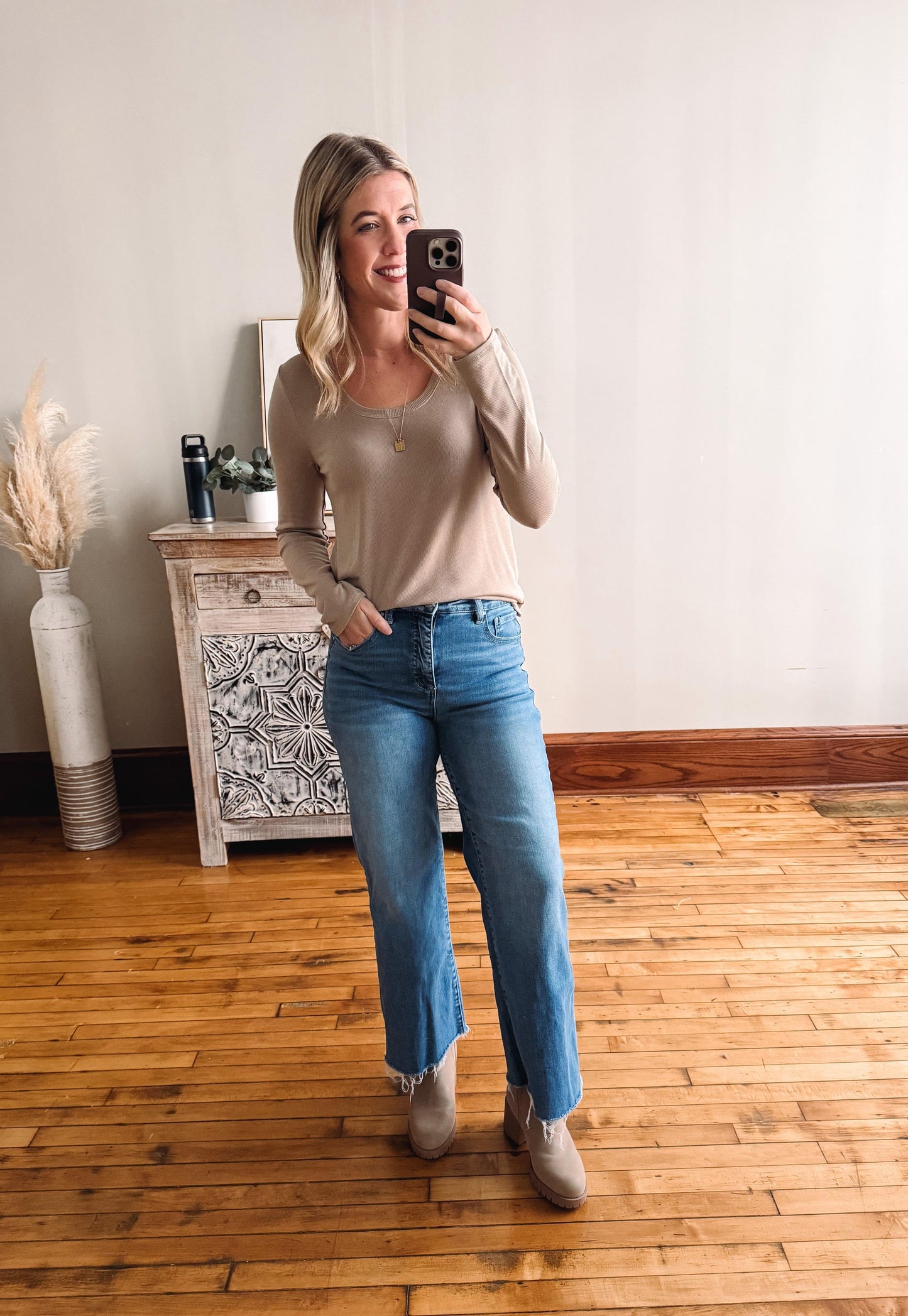 Woman taking a mirror selfie wearing a beige top and blue jeans in a room with wooden flooring.
