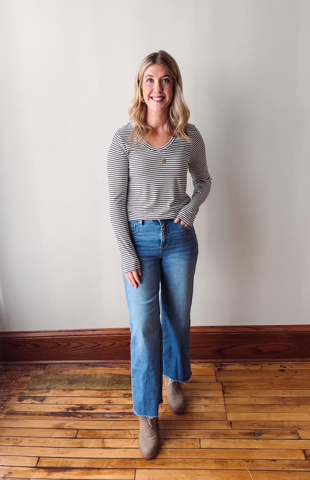 Woman wearing a striped shirt and jeans standing on a wooden floor with a white wall background