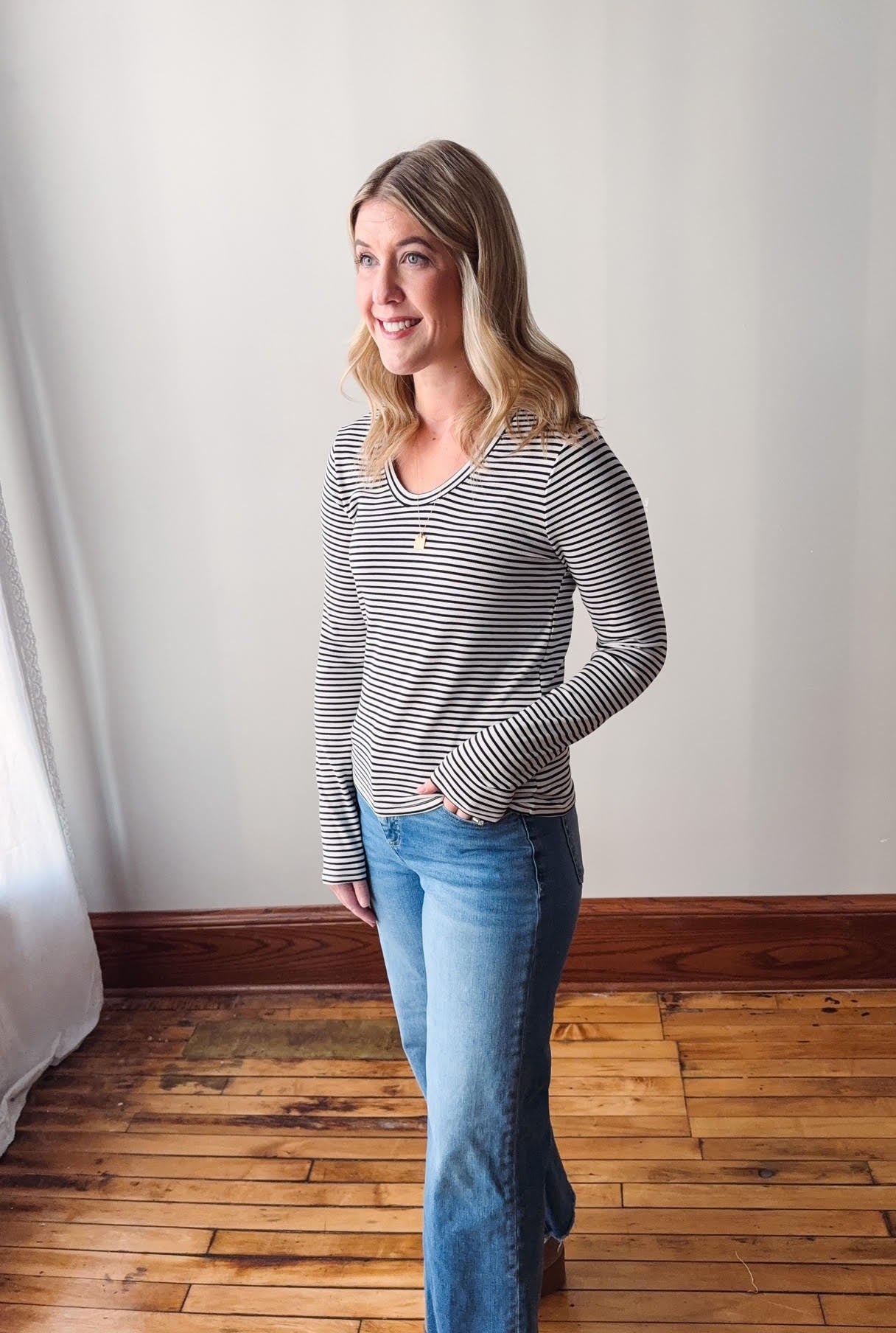 Woman wearing a striped shirt and jeans standing in a room with wooden flooring and white walls.