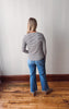 Person wearing a striped shirt and blue jeans standing on a wooden floor against a white wall.