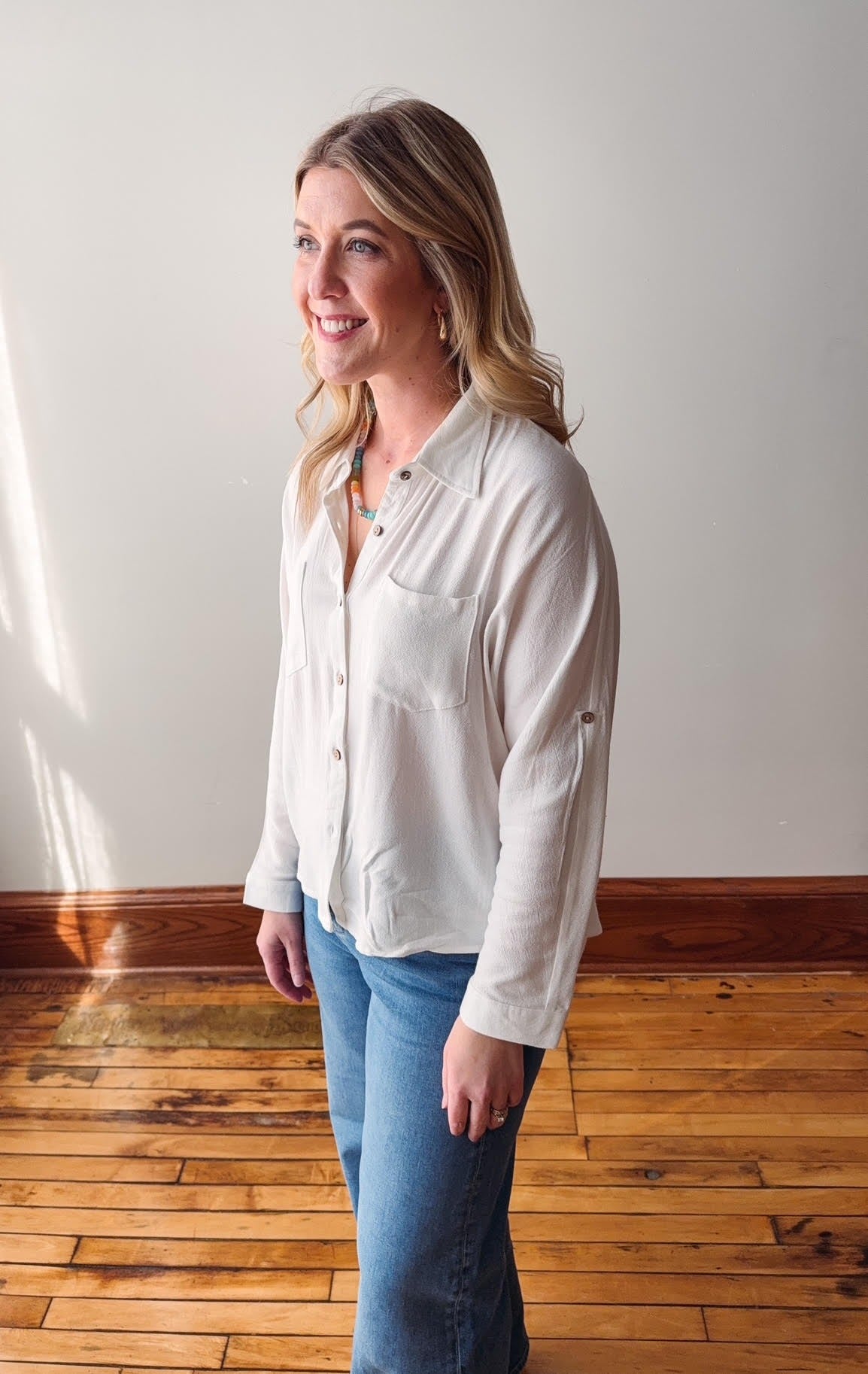 Woman wearing a white blouse and blue jeans standing in a room with wooden flooring and a white wall.
