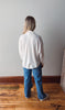 Person wearing a white shirt and blue jeans standing in a room with wooden flooring and a white wall.