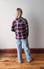 Person wearing a plaid shirt and jeans standing against a plain wall.