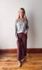 Woman wearing a light gray flannel shirt and burgundy pants standing on a wooden floor against a white wall.