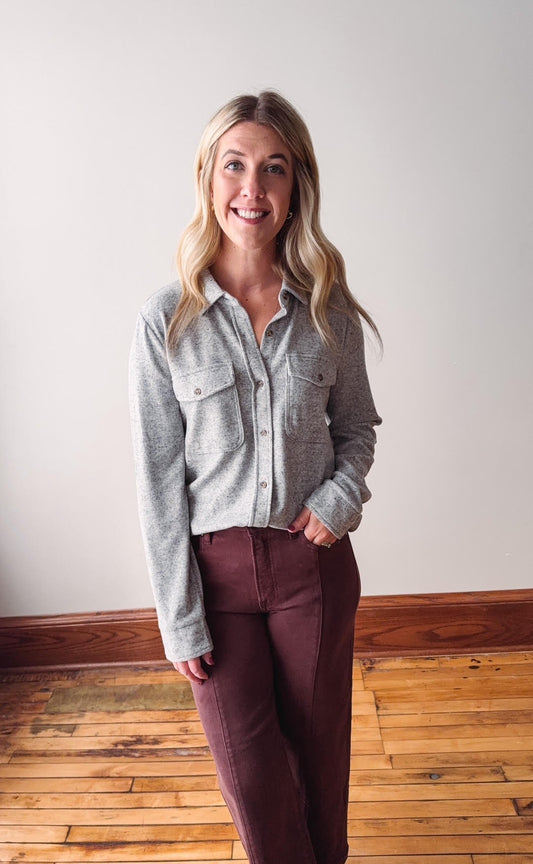 Woman wearing a gray button-up shirt and maroon pants standing in a room with wooden flooring.