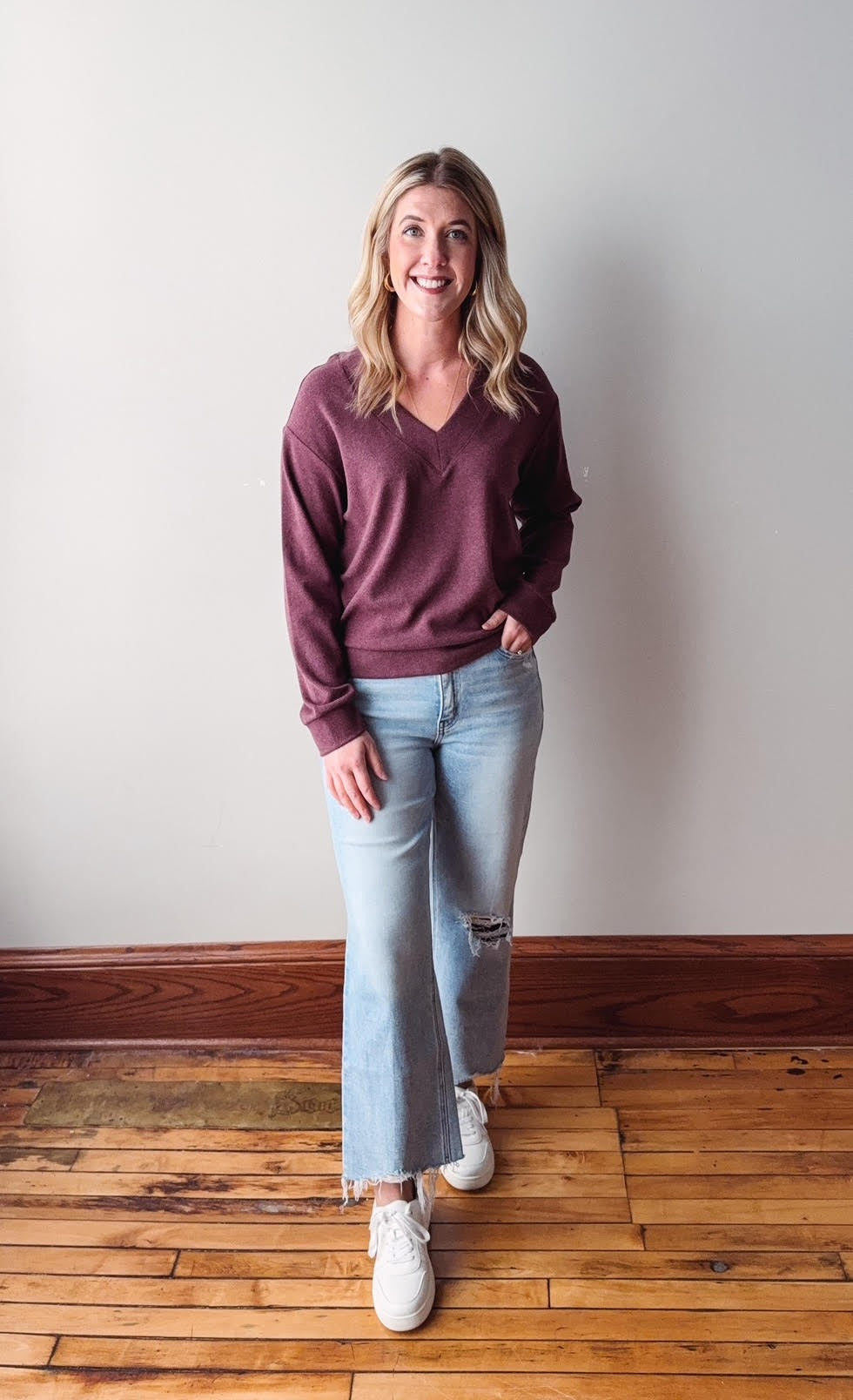 Woman wearing a maroon sweater and light blue jeans standing against a plain wall.