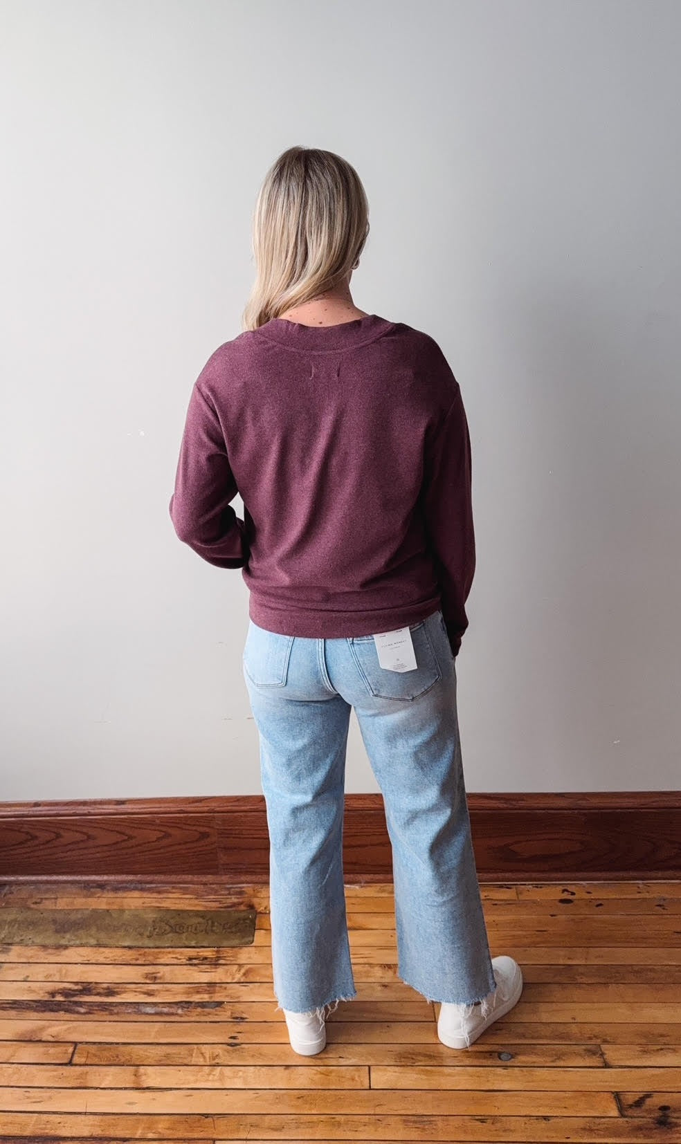 Person wearing a maroon sweatshirt and light blue jeans standing on a wooden floor with a white wall background.