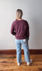 Person wearing a maroon sweatshirt and light blue jeans standing on a wooden floor with a white wall background.