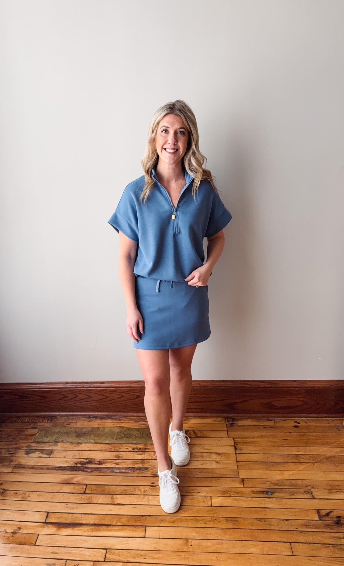 Woman wearing a blue outfit standing on a wooden floor against a white wall