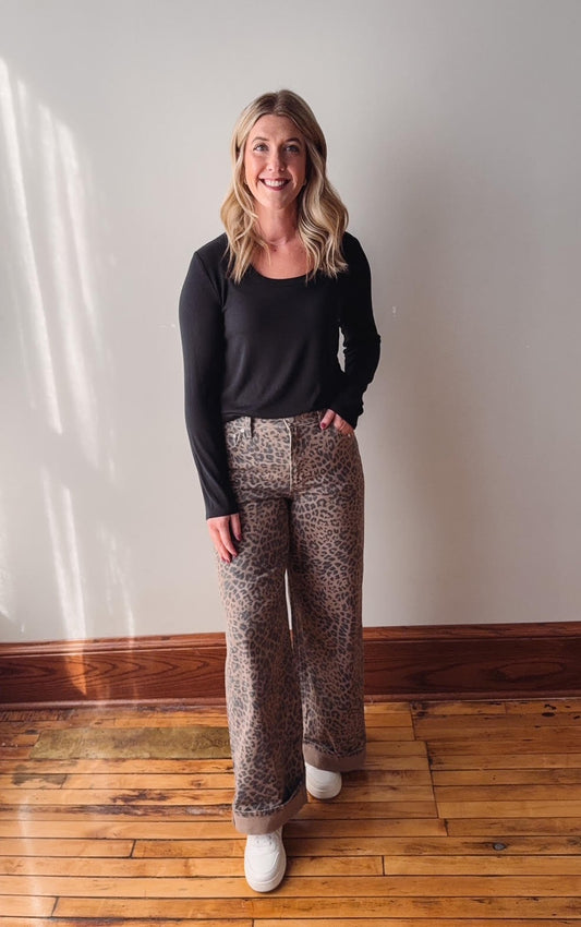 Woman wearing a black top and patterned pants standing in a room with wooden flooring and white walls.