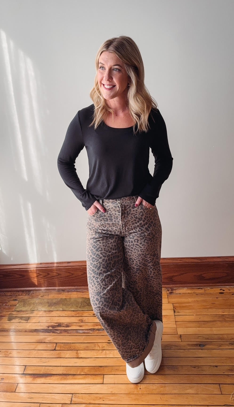 Woman wearing a black top and patterned pants standing on a wooden floor.