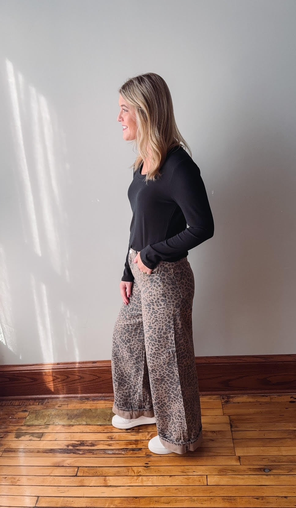 Woman wearing a black top and patterned pants standing against a plain wall.