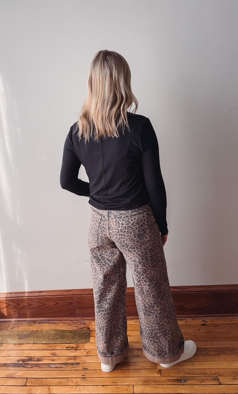 Person wearing a black long-sleeve top and patterned pants standing against a white wall.