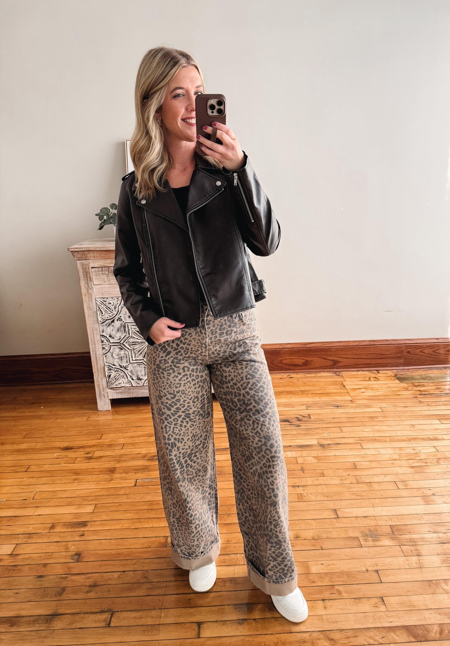 Woman taking a mirror selfie wearing a black leather jacket and patterned pants in a room with wooden flooring.