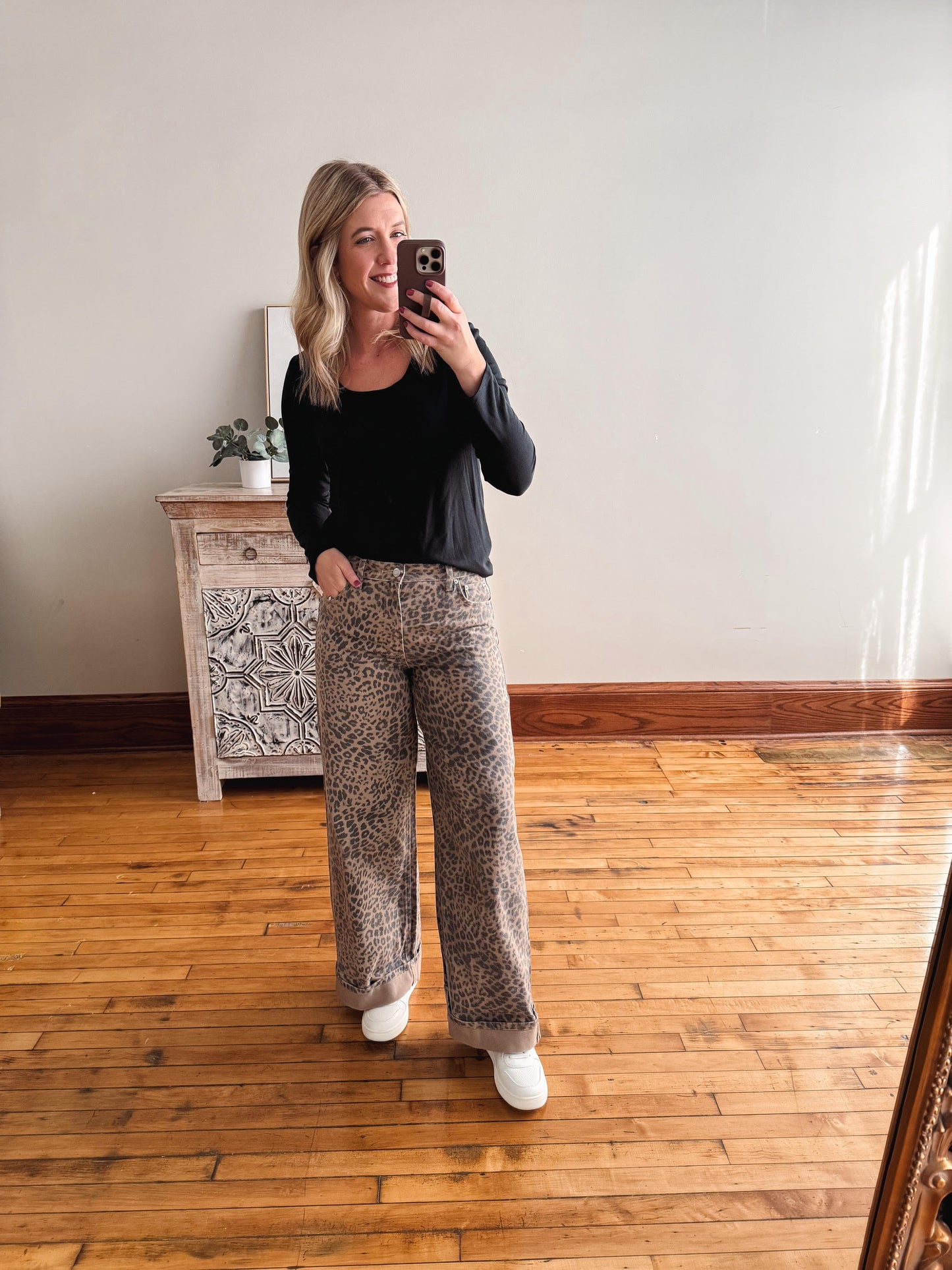 Woman taking a mirror selfie wearing a black top and patterned pants in a room with wooden flooring.