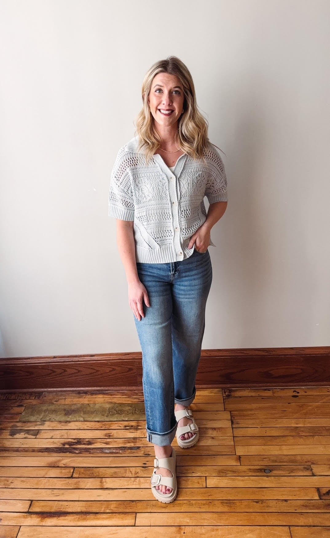 Woman wearing a white knit cardigan and blue jeans standing on a wooden floor.