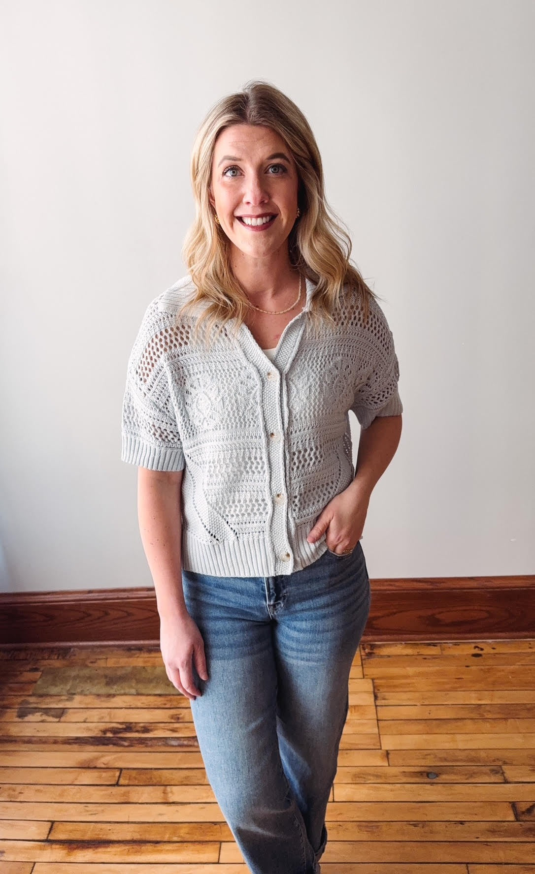 Woman wearing a white lace cardigan and blue jeans standing in a room with wooden flooring.