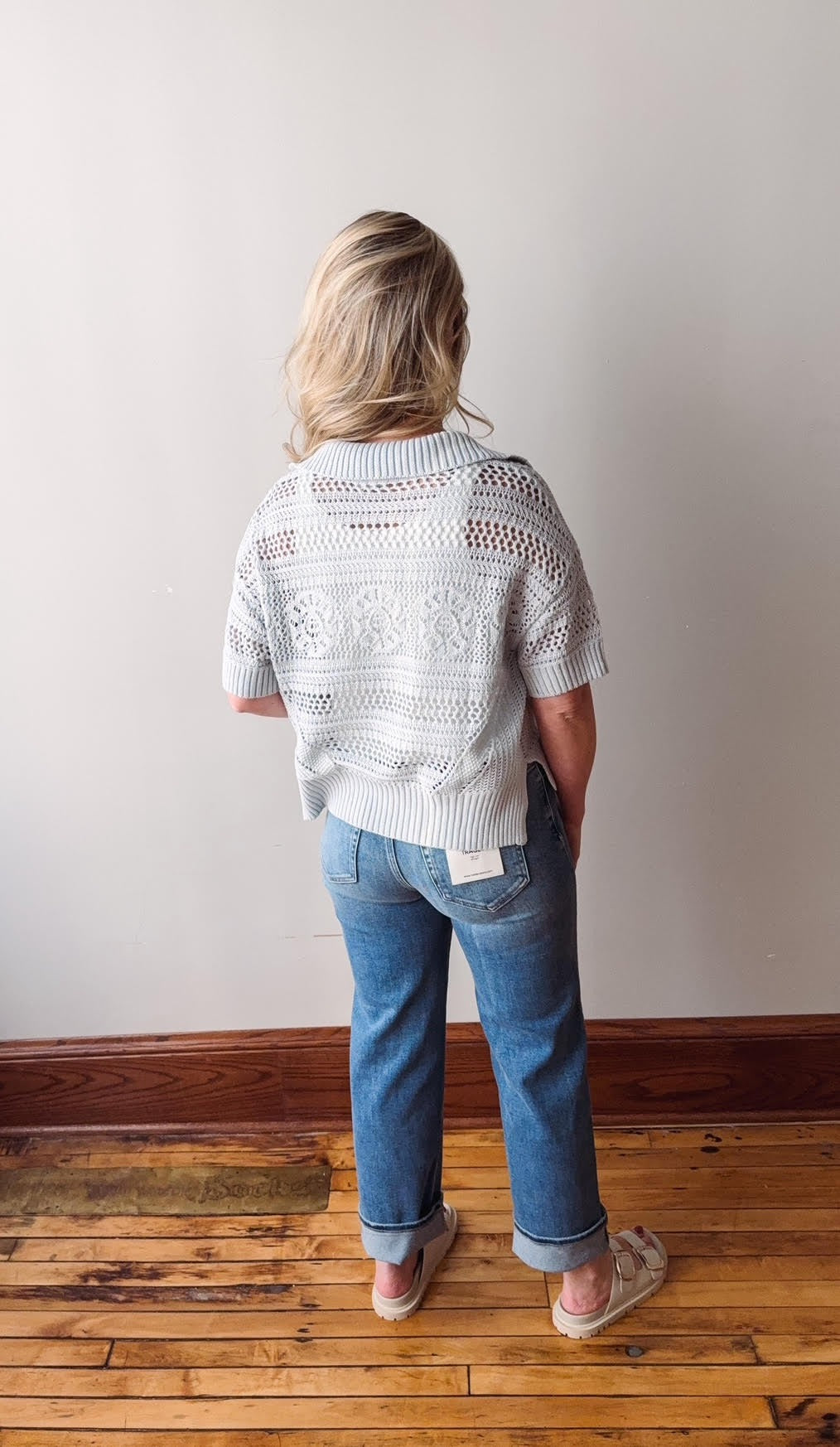 Person wearing a light gray knitted sweater and blue jeans standing against a plain white wall.