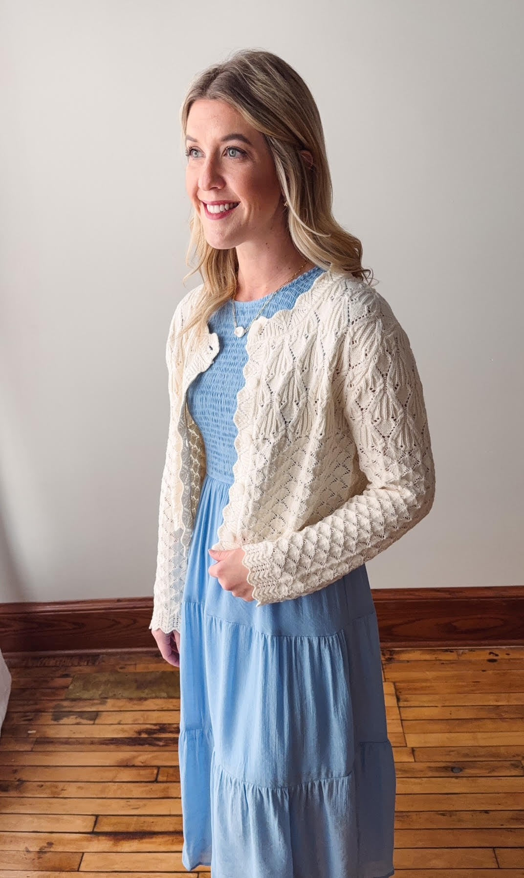 Woman wearing a white cable knit cardigan over a light blue dress on a wooden floor.