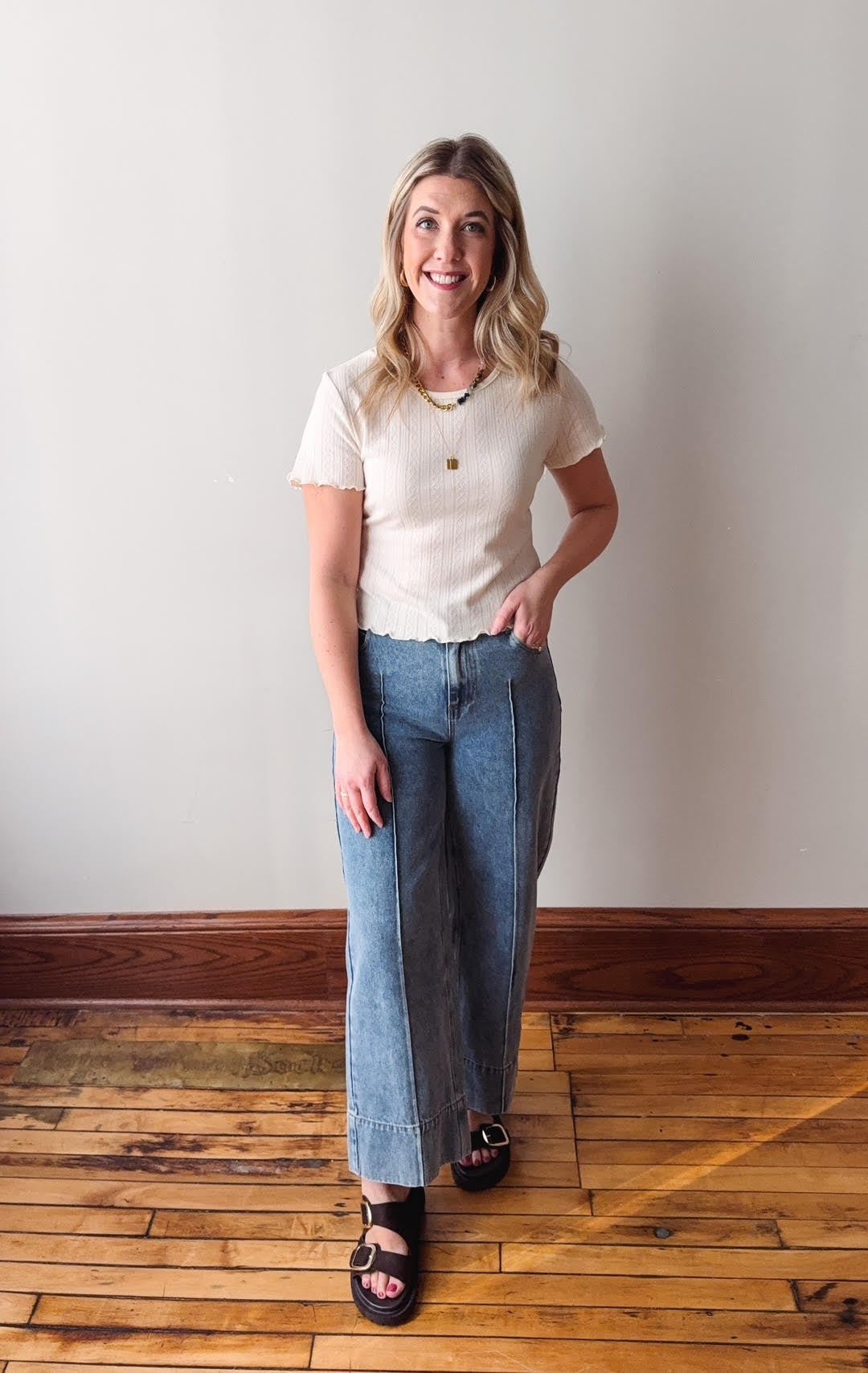 Woman wearing a white t-shirt and blue jeans standing on a wooden floor with a white wall background