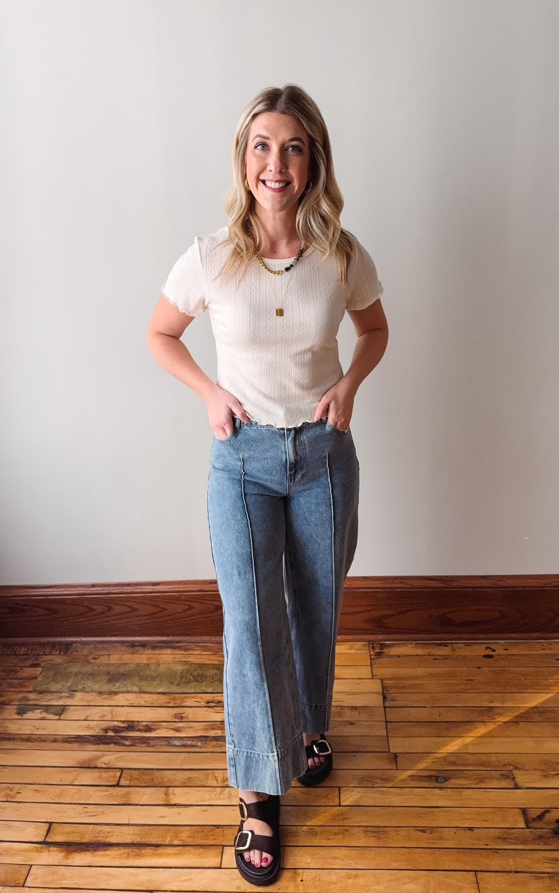 Woman wearing a white t-shirt and blue jeans standing on a wooden floor with a plain wall background