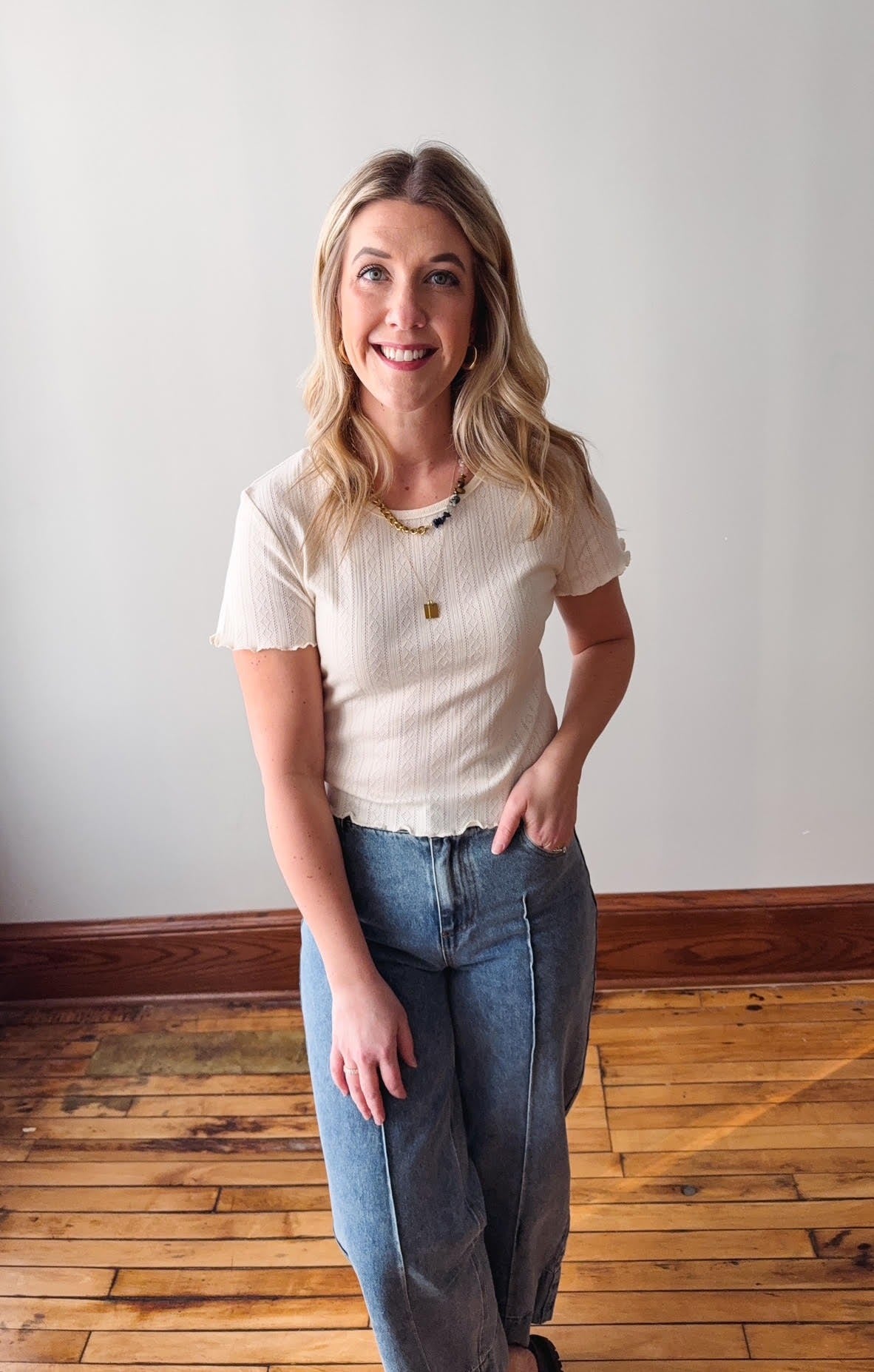 Woman wearing a white t-shirt and blue jeans standing on a wooden floor with a white wall background