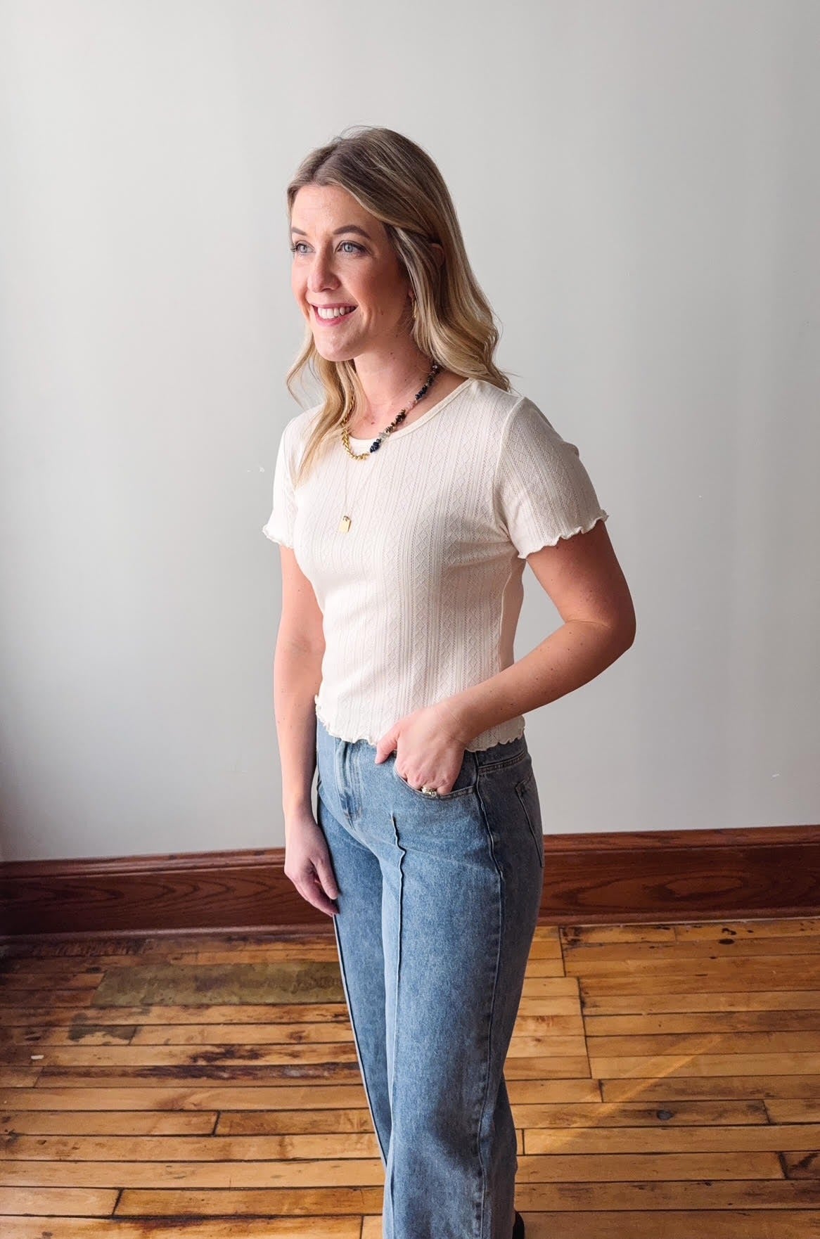 Woman wearing a white blouse and blue jeans standing in a room with wooden flooring and a white wall.